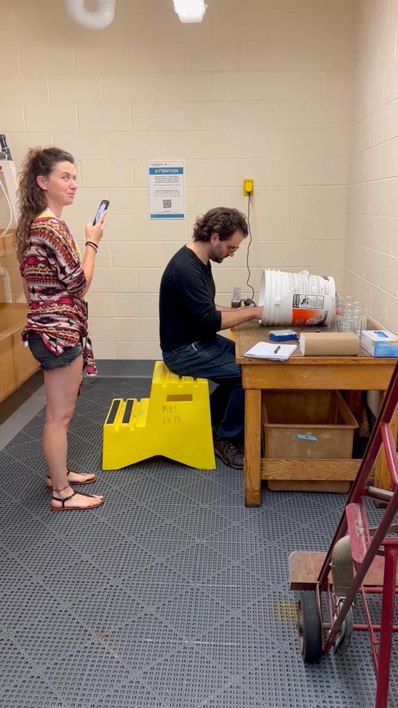A woman stands behind a man who is sitting at a desk flipping through a book.